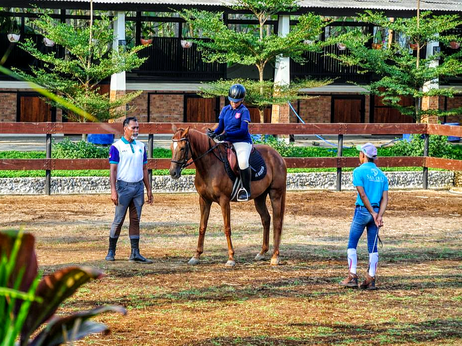 Seorang peserta latihan berkuda didampingi instruktur saat menunggang kuda di arena outdoor
WYN Stable